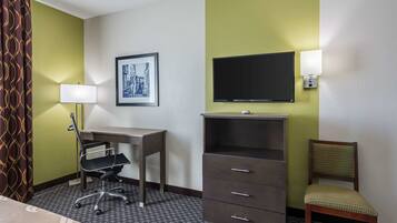 A hotel room with a desk, chair, TV, and a wall-mounted lamp.