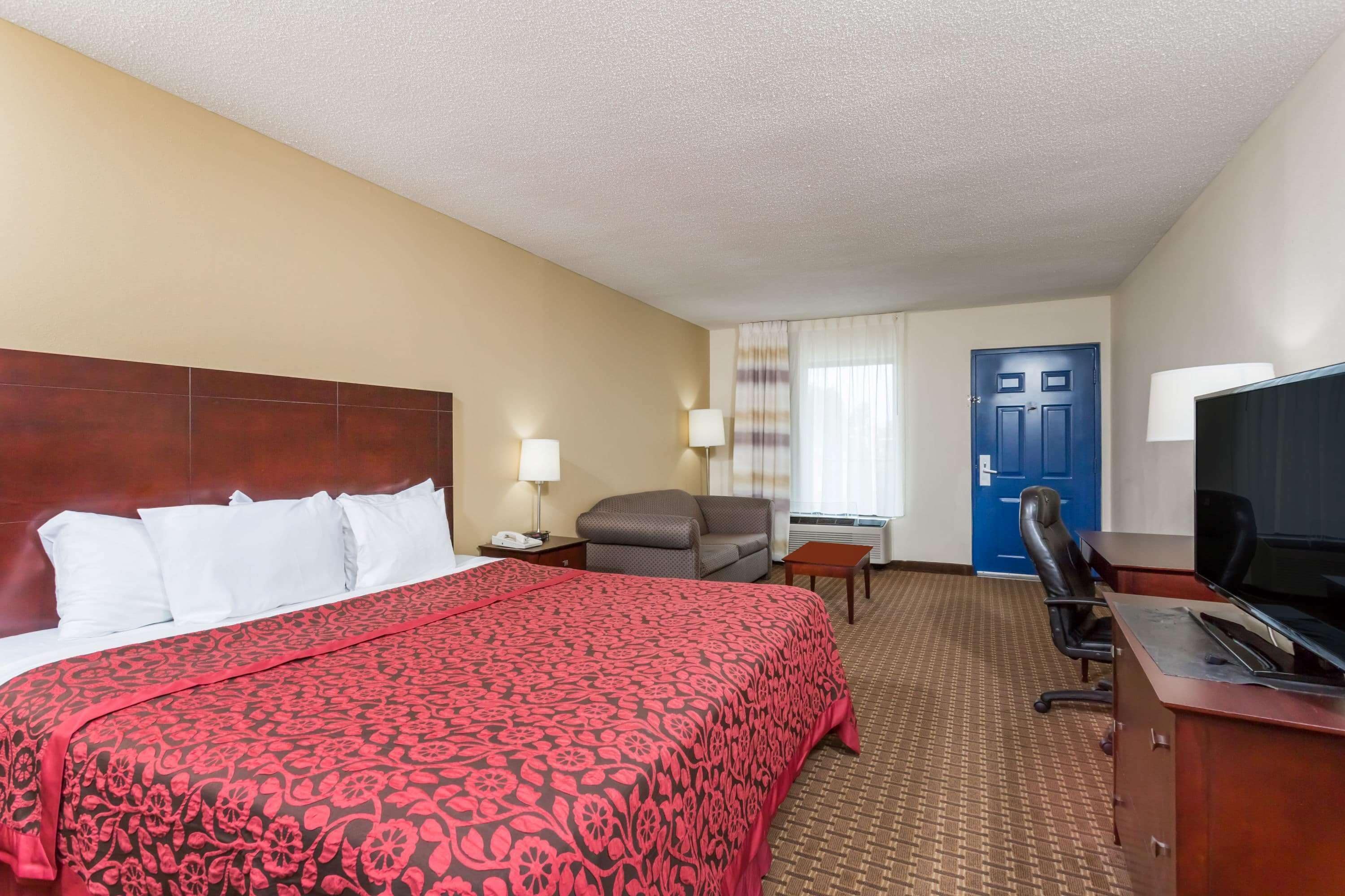 A hotel room with a large bed, a desk with a television, a chair, and a blue door.