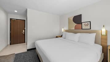 A hotel room with a bed, a nightstand, a wall-mounted lamp, a door, and a small plant.