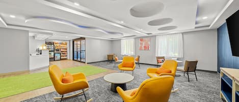 A modern interior with a reception desk, seating area with yellow chairs, a round table, and a TV screen.