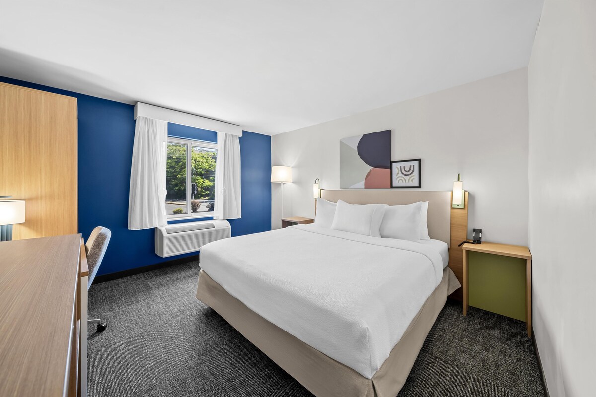 Une chambre d’hôtel avec un lit, un bureau, une fenêtre avec des rideaux, une lampe fixée au mur et un tableau encadré.
