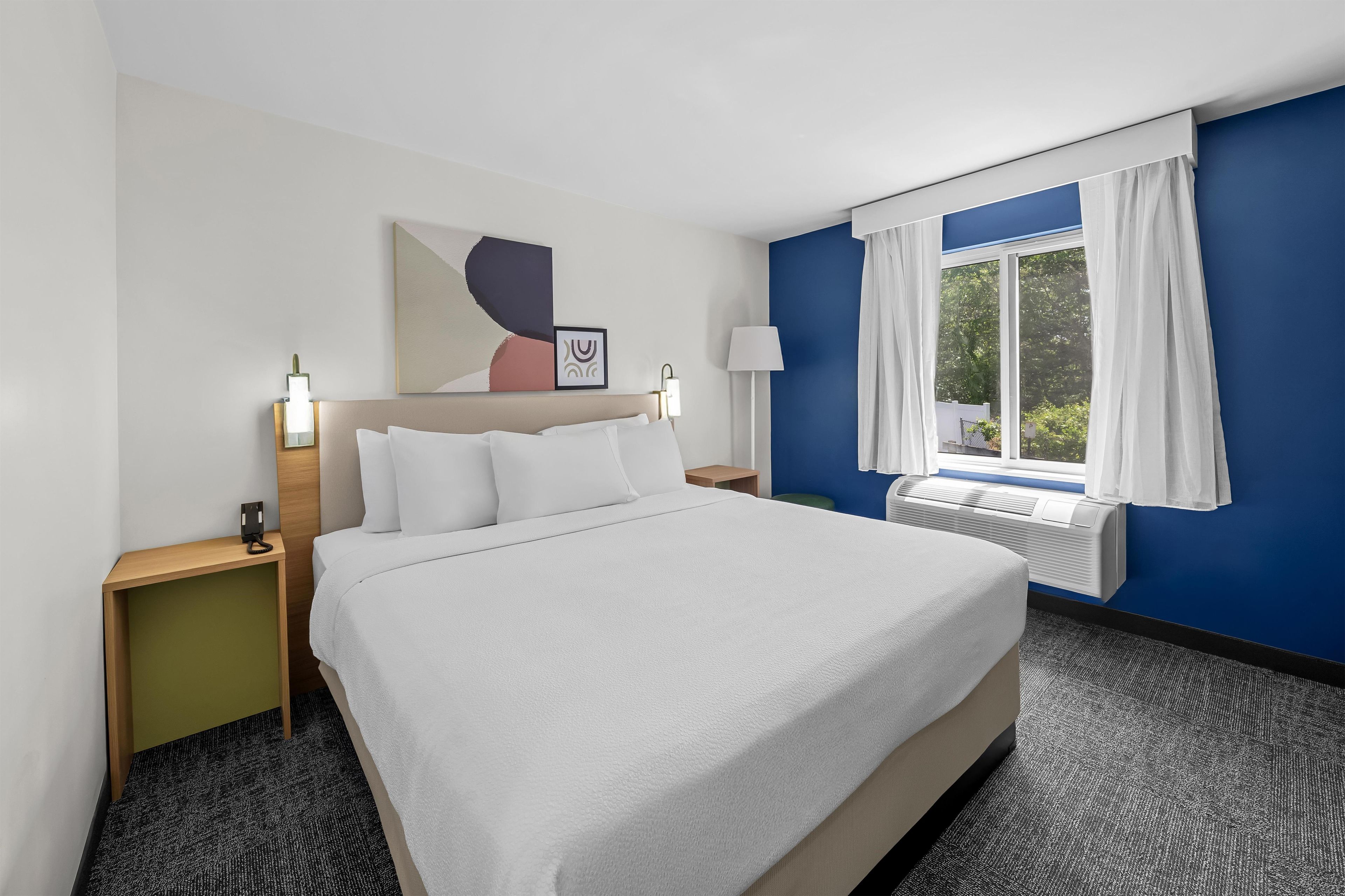A hotel room with a large bed, a nightstand, a lamp, a window with curtains, and a wall-mounted air conditioner.