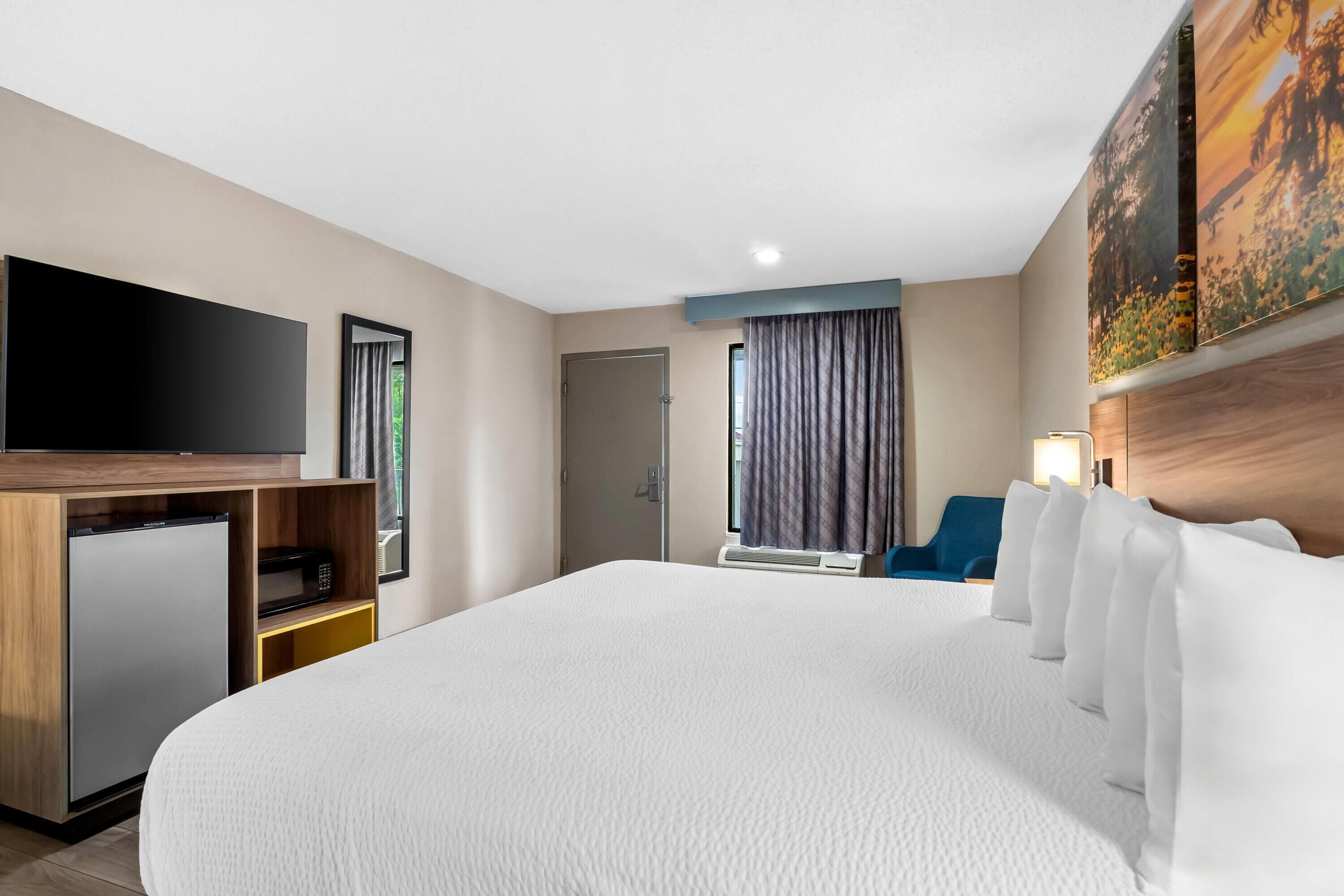 Habitación de hotel con una cama grande, televisión de pantalla plana, un refrigerador pequeño, escritorio y ventana con cortinas.