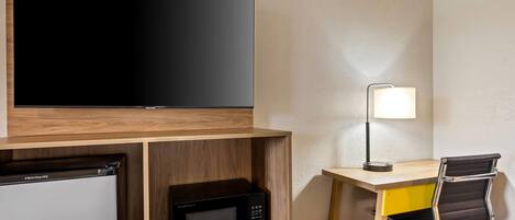 Ein kleines Hotelzimmer mit Flachbildfernseher, Mikrowelle, kleinem Kühlschrank, Schreibtisch mit Stuhl und Nachttischlampe.