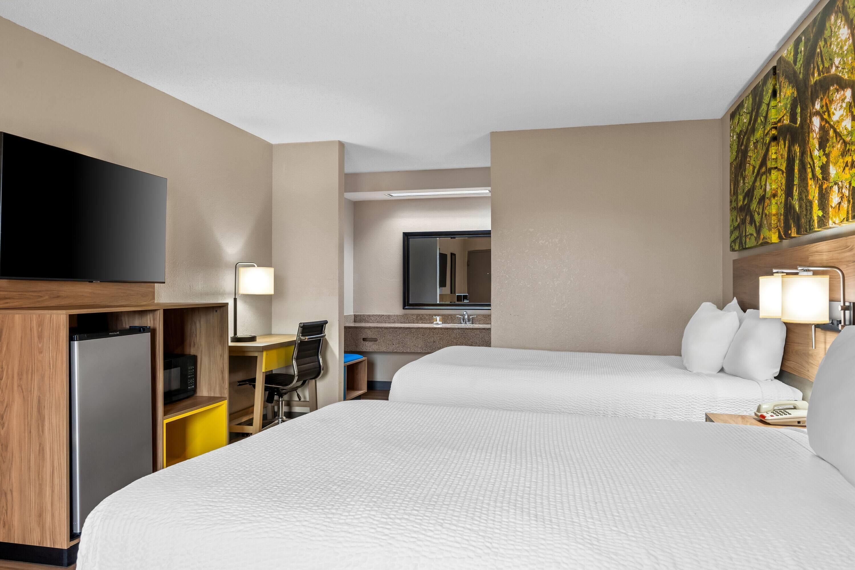 A hotel room with two beds, a desk with a chair, a television, a refrigerator, a microwave, a sink, and a painting on the wall.