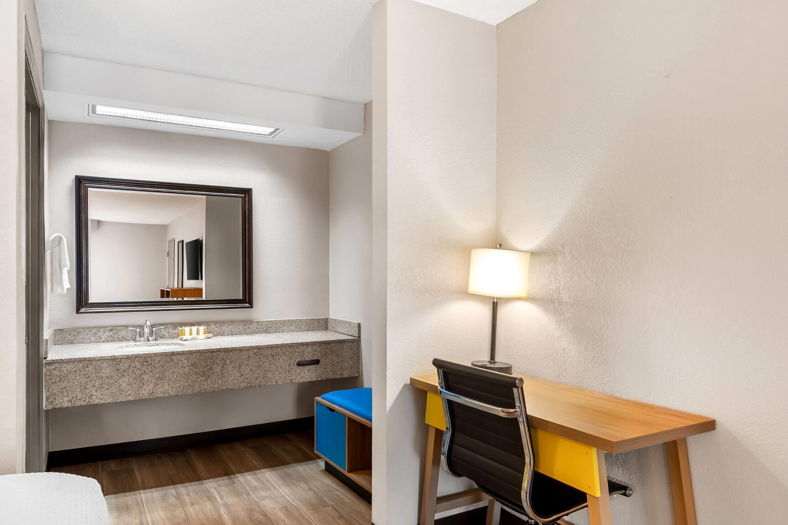 Une chambre d’hôtel dotée d’une salle de bain avec un lavabo au comptoir en granit, un miroir et un bac de rangement bleu. On y trouve également un bureau avec une chaise et une lampe.