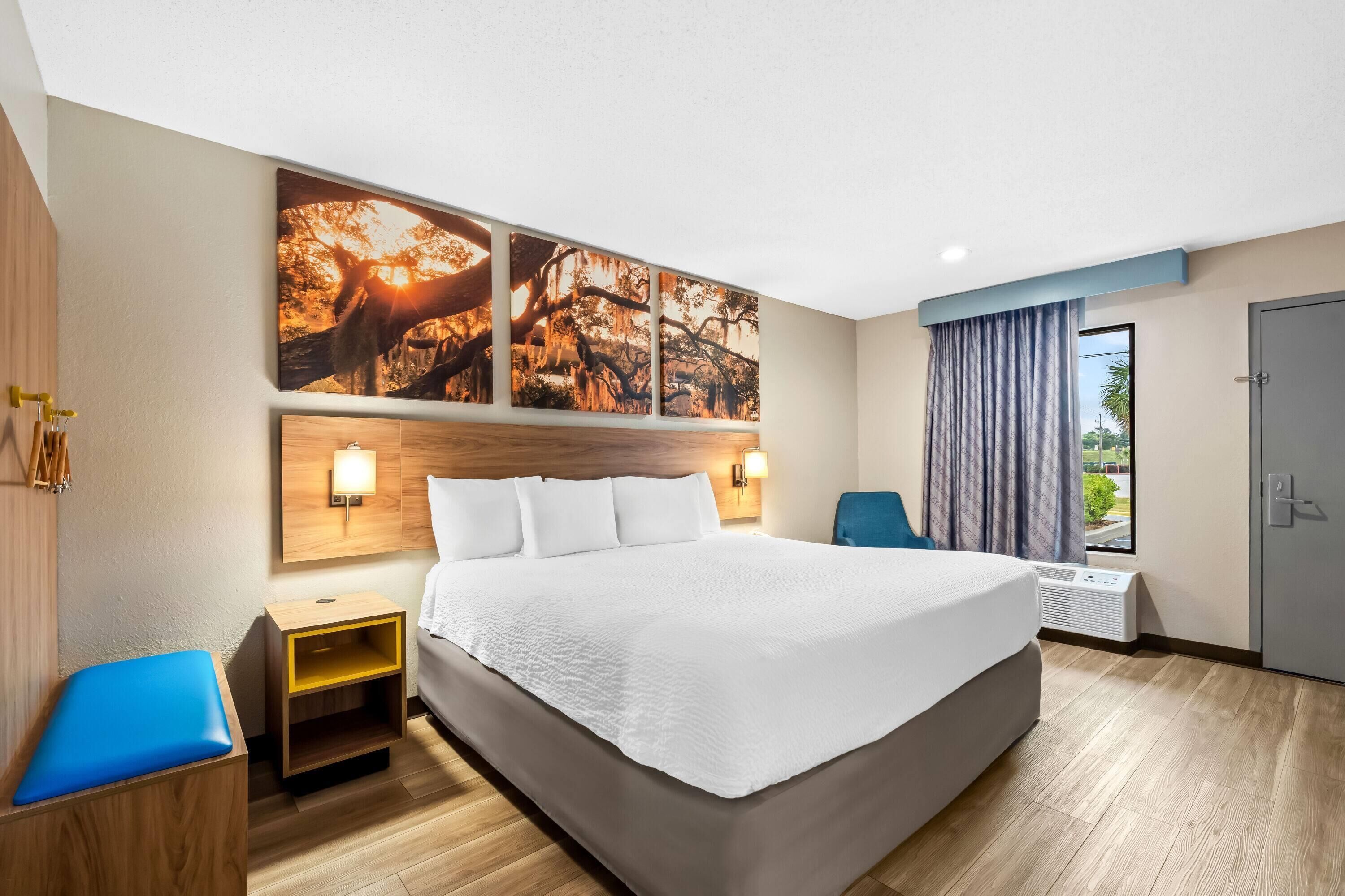 Habitación de hotel con una cama grande, una mesita de noche, una silla azul, un televisor instalado en la pared y una ventana con cortinas.