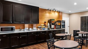 Eine moderne Küche mit dunklen Schränken, einer Mikrowelle, einer Kaffeemaschine und einem Kühlschrank. Runde Tische und Stühle bieten Platz zum Essen.