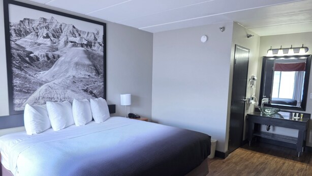Una habitación de hotel con una cama grande, una mesita de noche con lámpara, un baño con espejo y un cuadro en la pared.