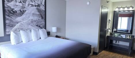 Een hotelkamer met een groot bed, een nachtkastje met een lamp, een badkamer met een spiegel en een lijst met een afbeelding aan de muur.