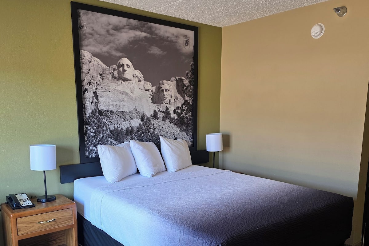 Una camera d'albergo con un letto, due comodini, un telefono a parete e un quadro grande con cornice.
