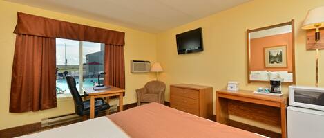 A hotel room with a bed, desk, chair, dresser, TV, and a view of a pool.