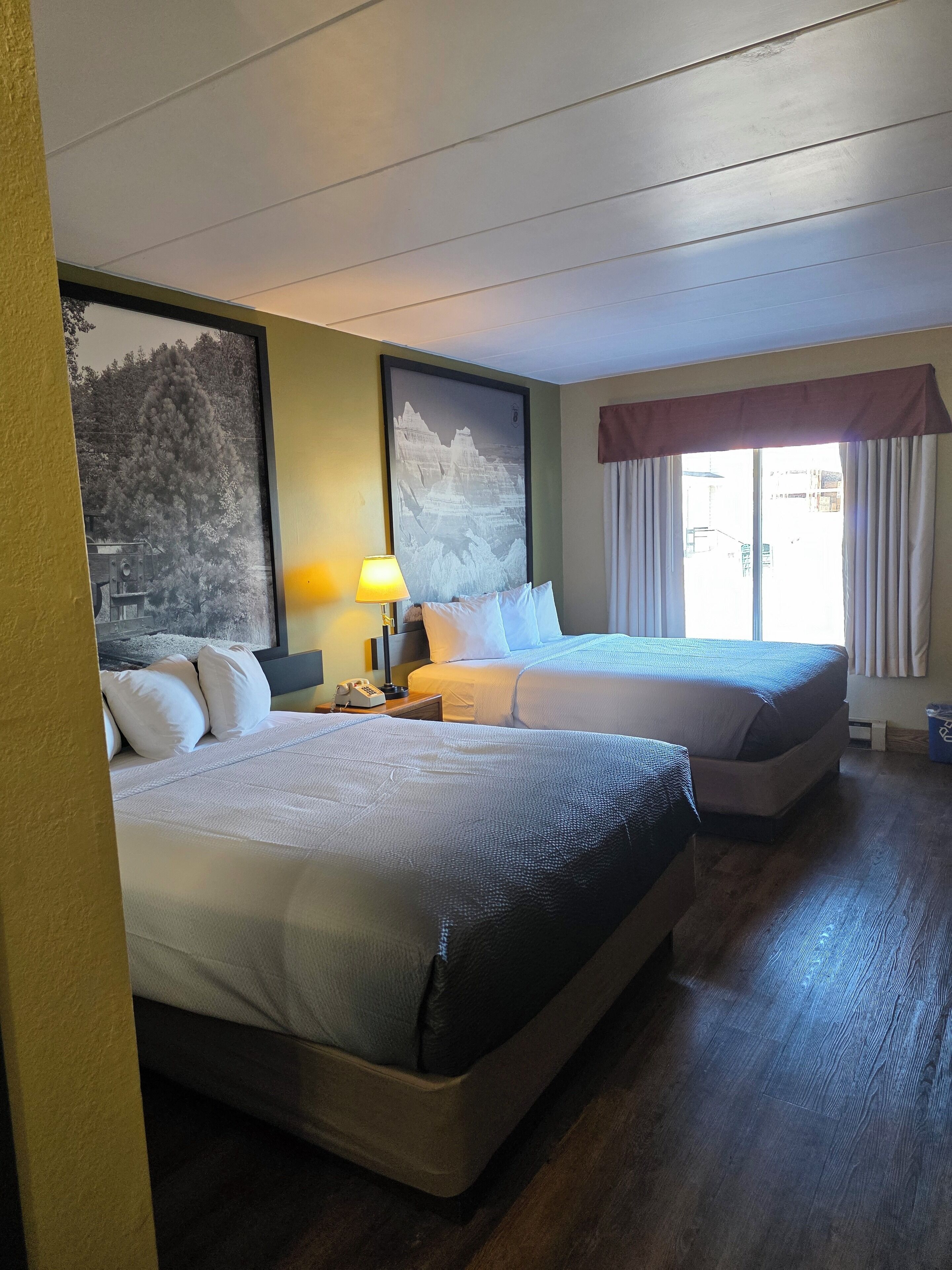 Een hotelkamer met twee bedden, een groot gepresenteerd schilderij van een berglandschap, een bedlampje en een raam met gordijnen.