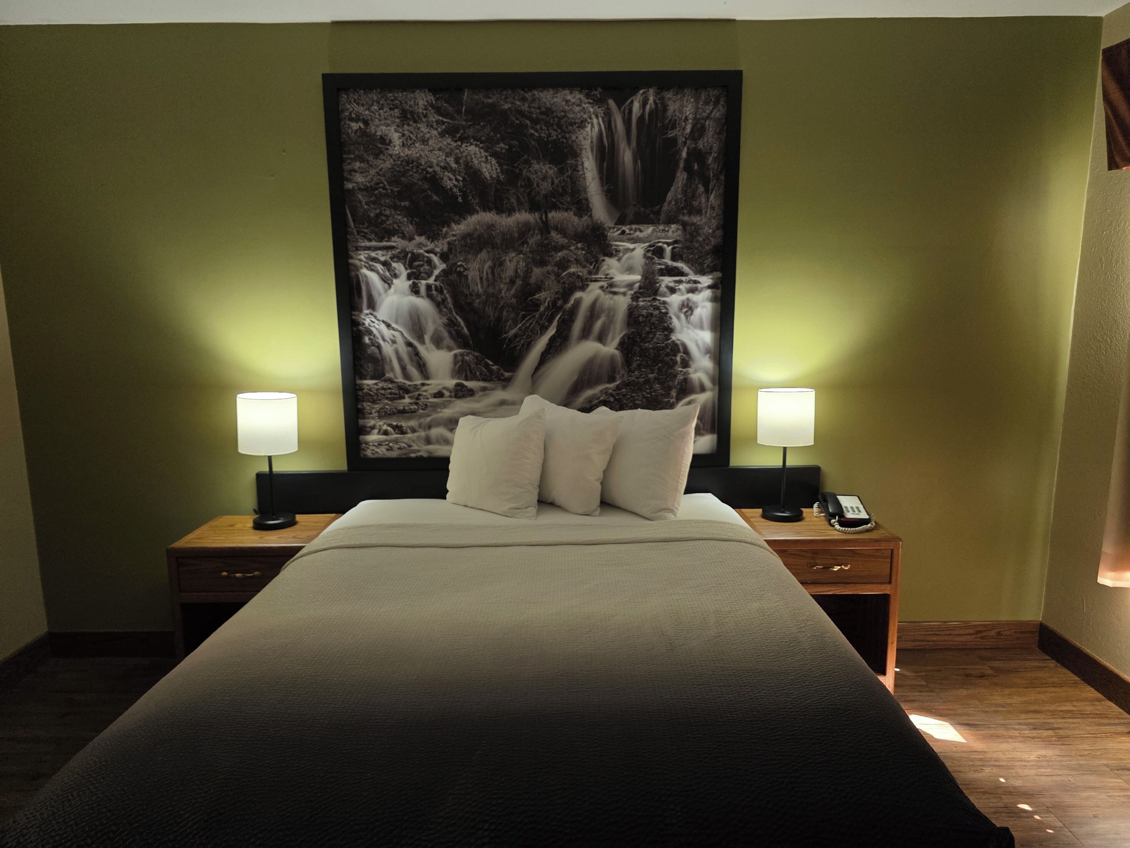 A hotel room with a large bed, two bedside lamps, a nightstand with a phone, and a large waterfall painting on the wall.