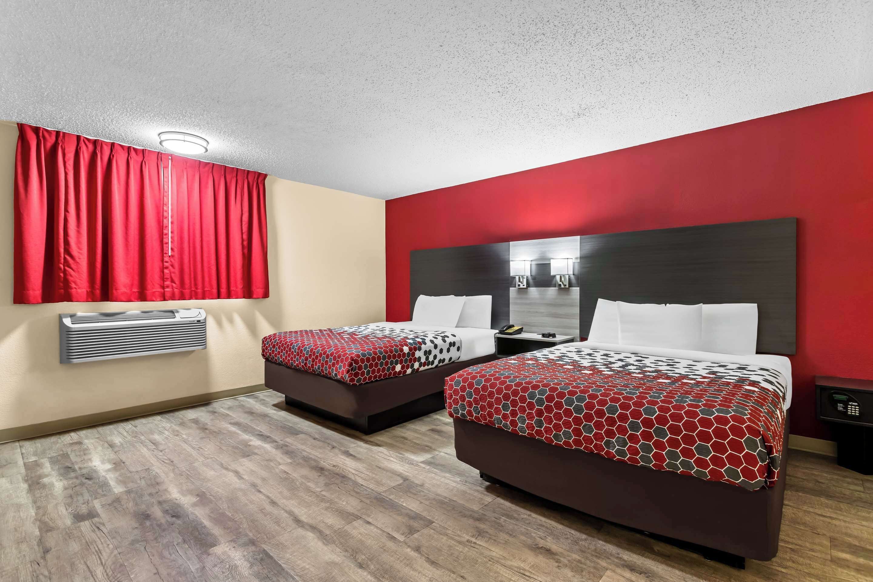 A hotel room with two beds, red curtains, a red and beige wall, a wooden floor, and a modern headboard.