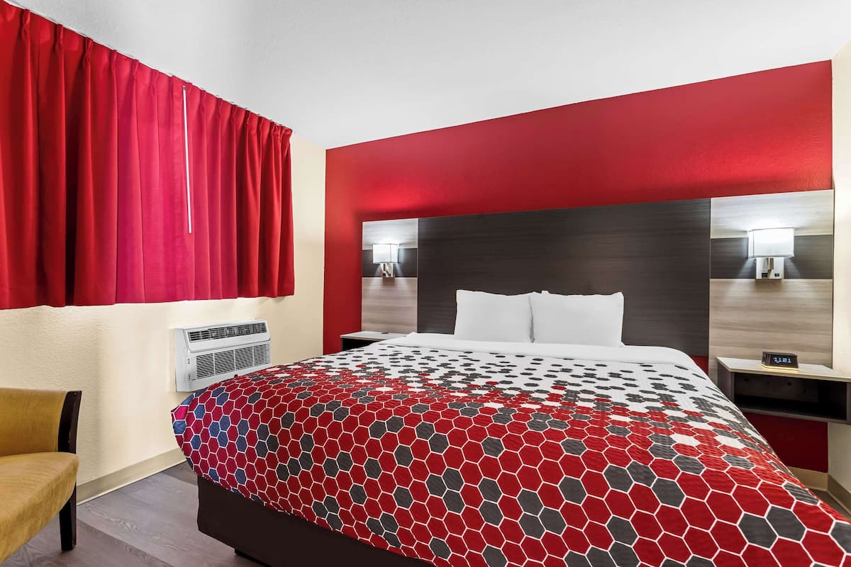 A hotel room with a bed, a nightstand, a chair, a red curtain, and a wall-mounted air conditioner.