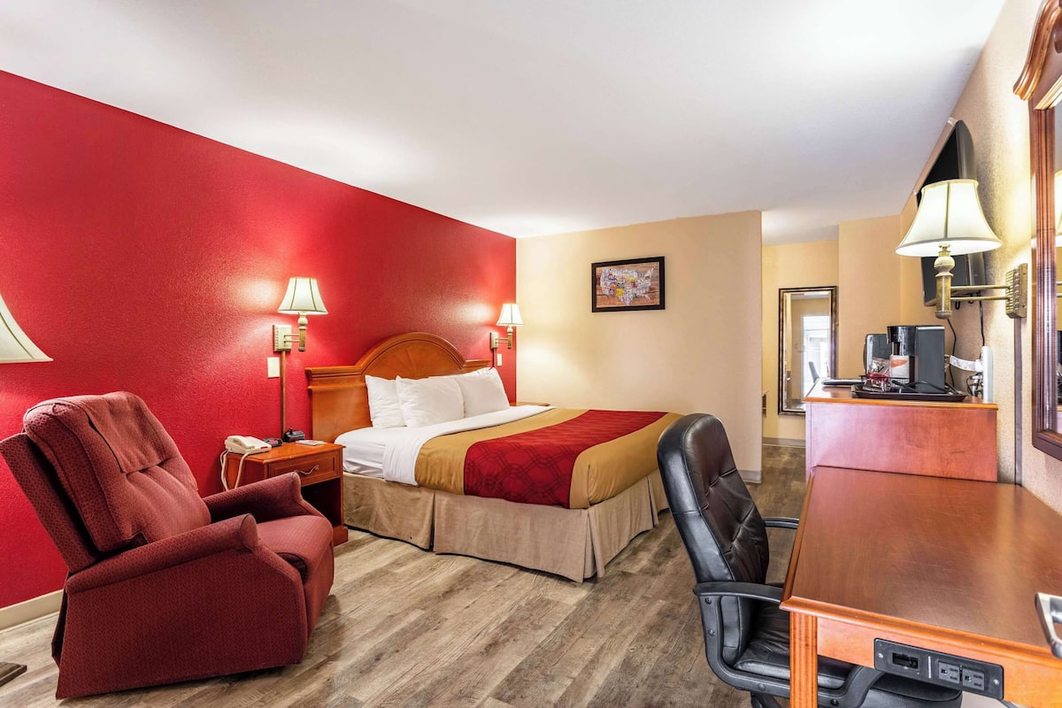 A hotel room with a bed, a red armchair, a desk with a computer, and a lamp.