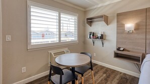 A small room with a round table, two chairs, a window with blinds, a wall-mounted shelf with a coffee maker, and a wall-mounted light. - Harbor House Inn Morro Bay (Morro Bay)
