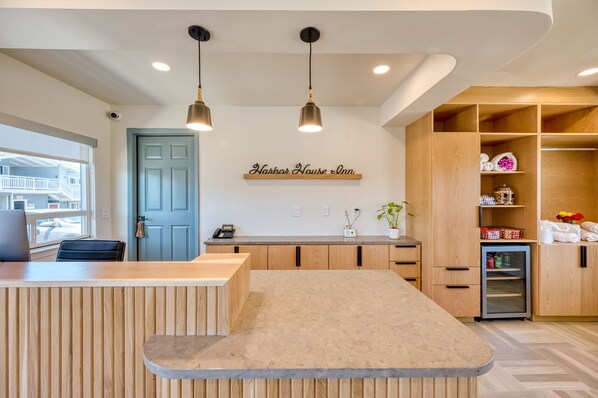 Un hall d’hôtel moderne avec un comptoir en bois, une porte bleue et une enseigne indiquant « Harbor House Inn ». On y trouve des lampes suspendues, un petit réfrigérateur et une plante posée sur le comptoir.