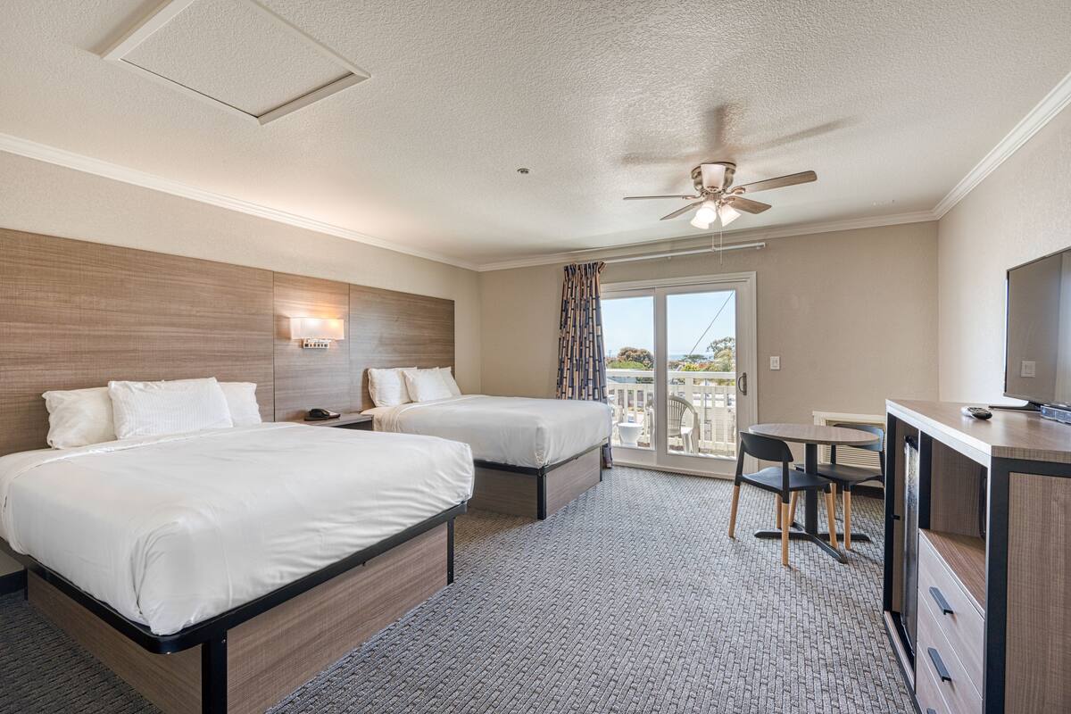 A hotel room with two beds, a desk, a chair, a ceiling fan, and a view of the outdoors.
