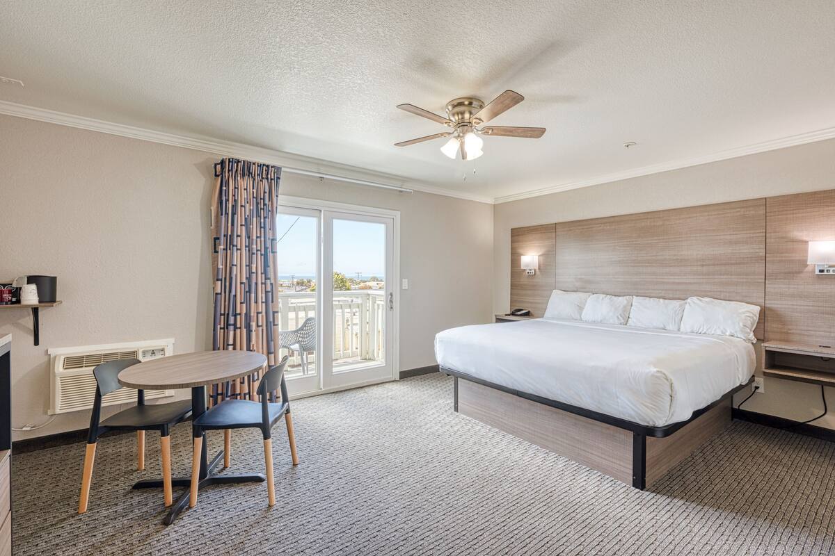 A hotel room with a bed, a ceiling fan, a table with chairs, and a view through the glass door.
