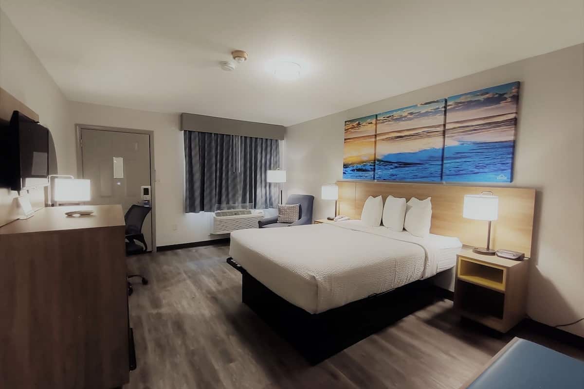 A hotel room with a bed, desk, chair, and artwork on the wall.
