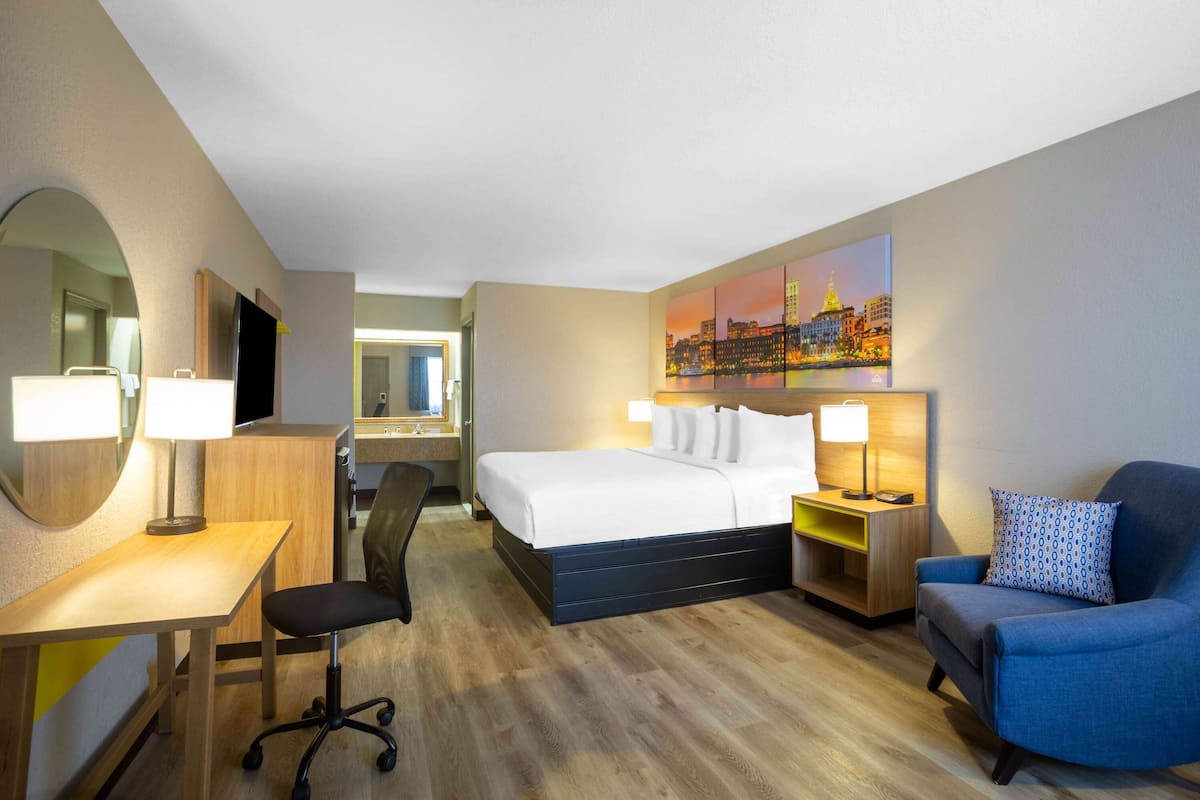 A hotel room with a bed, desk, chair, and a painting on the wall.
