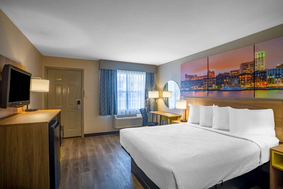 A hotel room with a bed, desk, TV, and a cityscape painting on the wall.