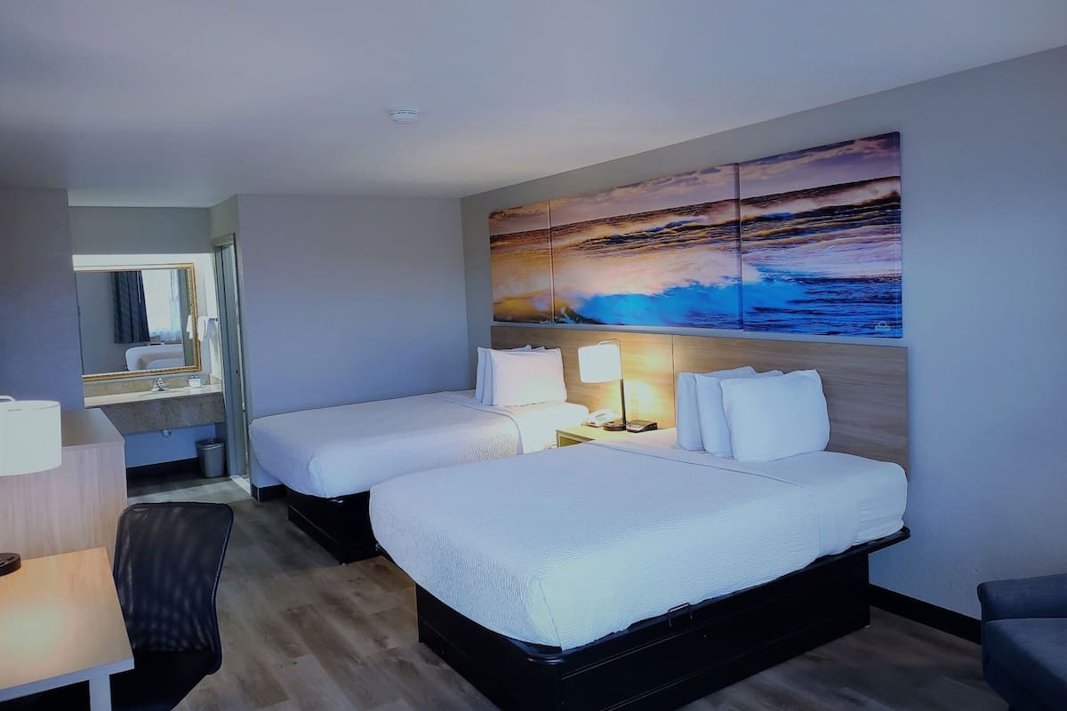 A hotel room with two beds, a desk, a chair, a bathroom with a mirror, and a large painting of a sunset over the ocean.