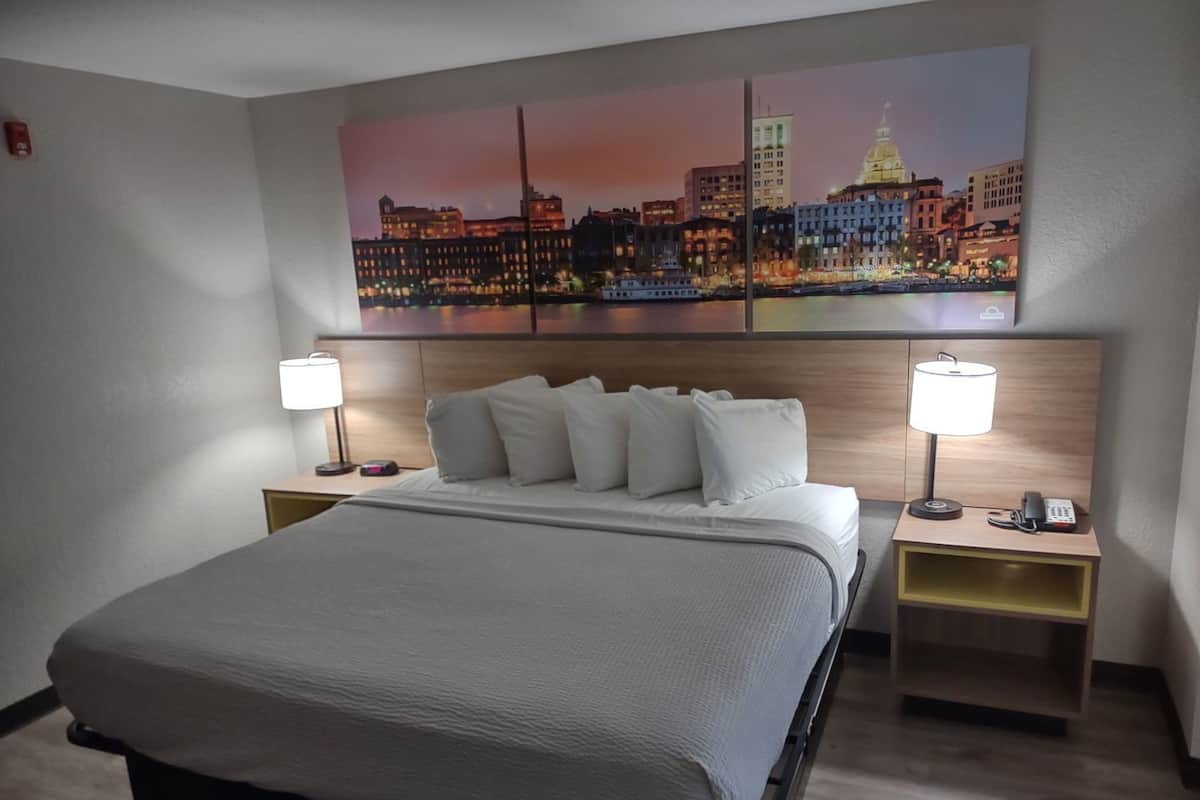 A hotel room with a bed, two bedside tables, a nightstand, a wall-mounted lamp, and a cityscape painting above the bed.