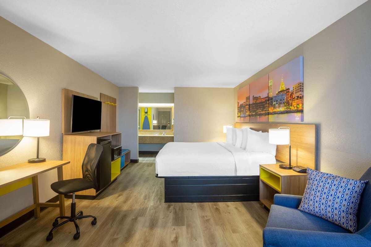 A hotel room with a bed, a desk with a chair, a TV, and a painting on the wall.