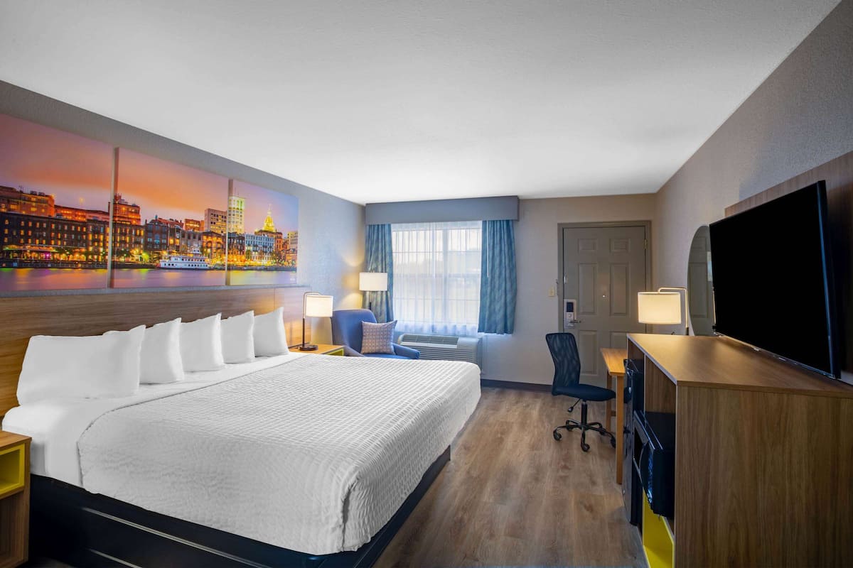 A hotel room with a large bed, a desk, a chair, a TV, and a cityscape painting on the wall.