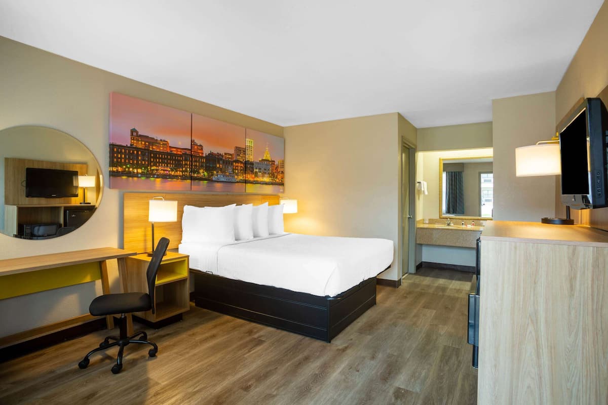 A hotel room with a bed, desk, chair, TV, and a painting of a cityscape.