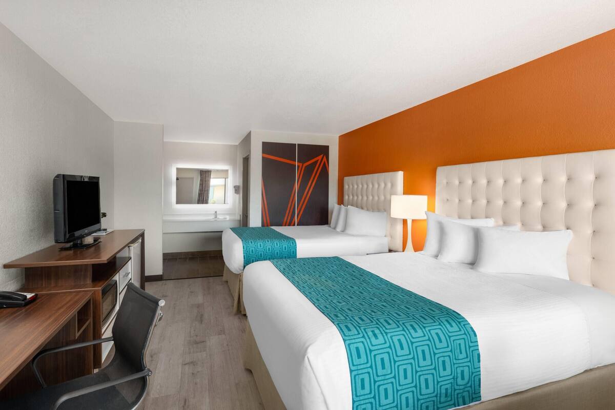 A hotel room with two beds, a desk with a computer, a TV, and a bathroom visible in the background.