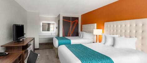 A hotel room with two beds, a desk with a computer, a TV, and a bathroom visible in the background.