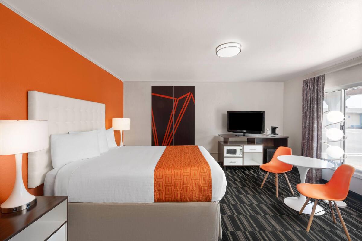 A hotel room with a bed, bedside lamps, a desk with a flat-screen TV, two orange chairs, and a large window with curtains.