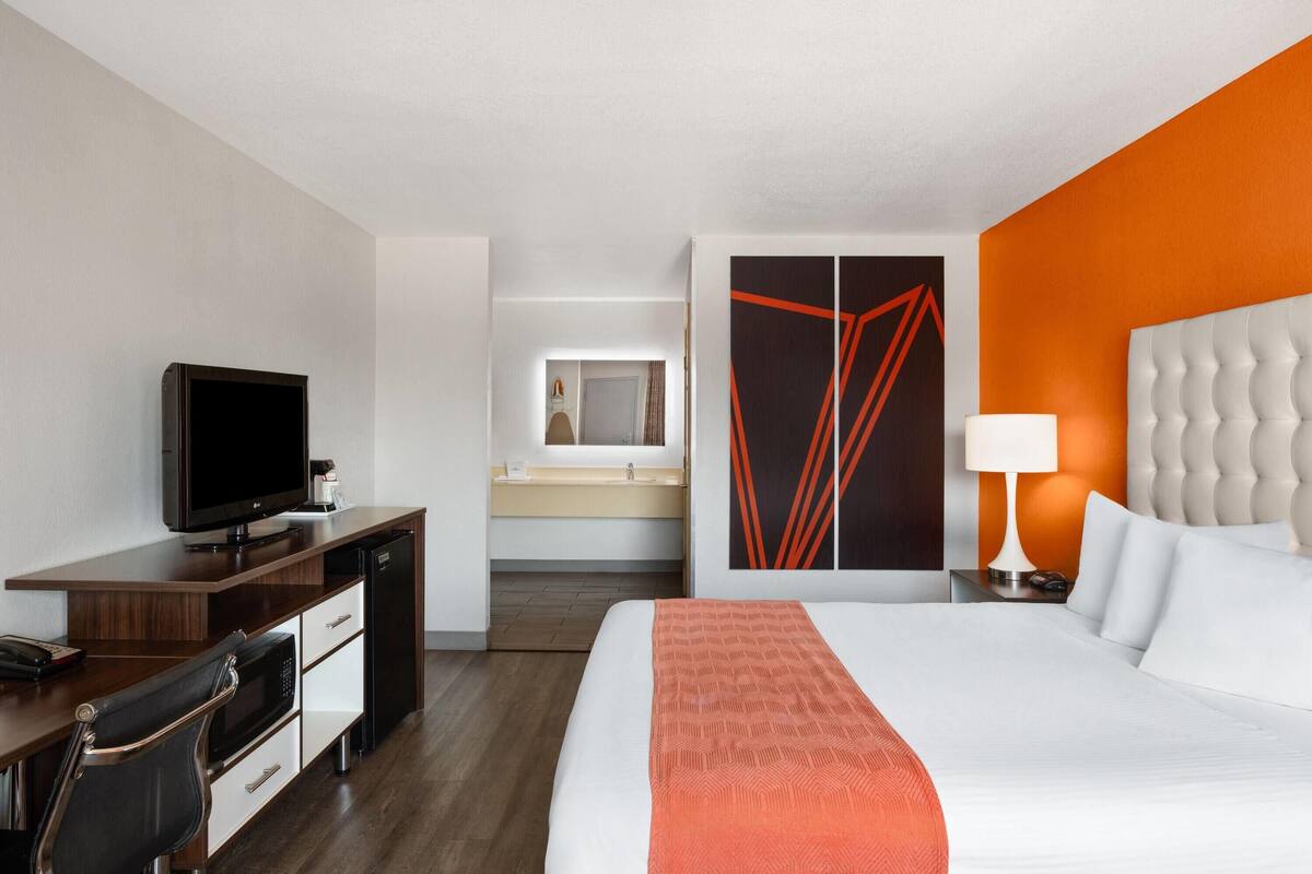 A hotel room with a bed, a desk with a television, a chair, a wardrobe, and a bathroom visible through an open door.