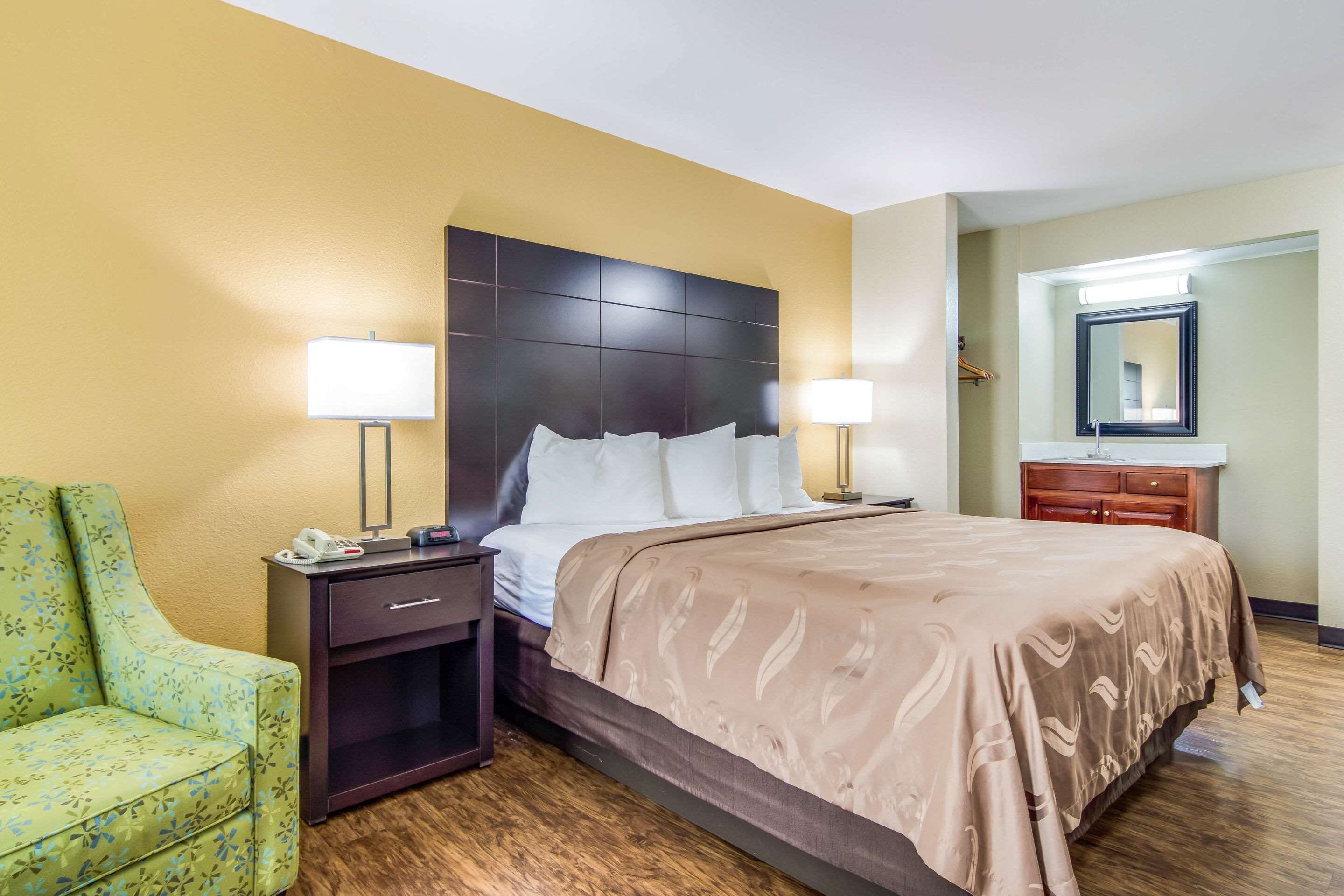 A hotel room with a large bed, bedside lamps, a nightstand, a green armchair, and a wooden floor.