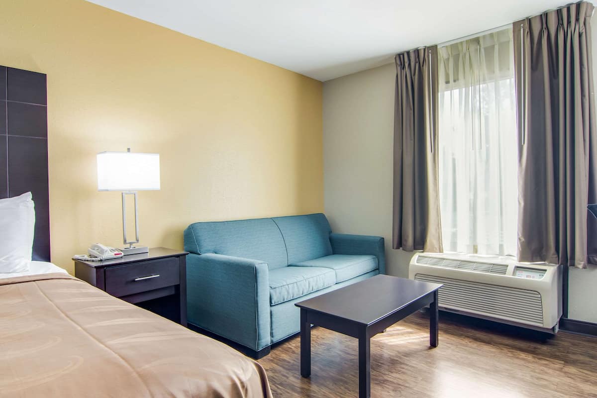 Quarto de hotel com cama, sofá, mesa pequena, abajur, aquecedor e janela com cortinas.