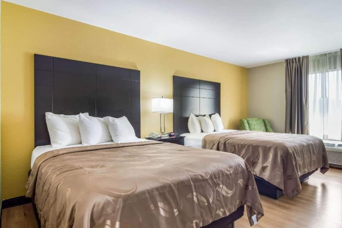 Quarto de hotel com duas camas, uma mesinha de cabeceira, uma luminária e uma janela com cortinas.