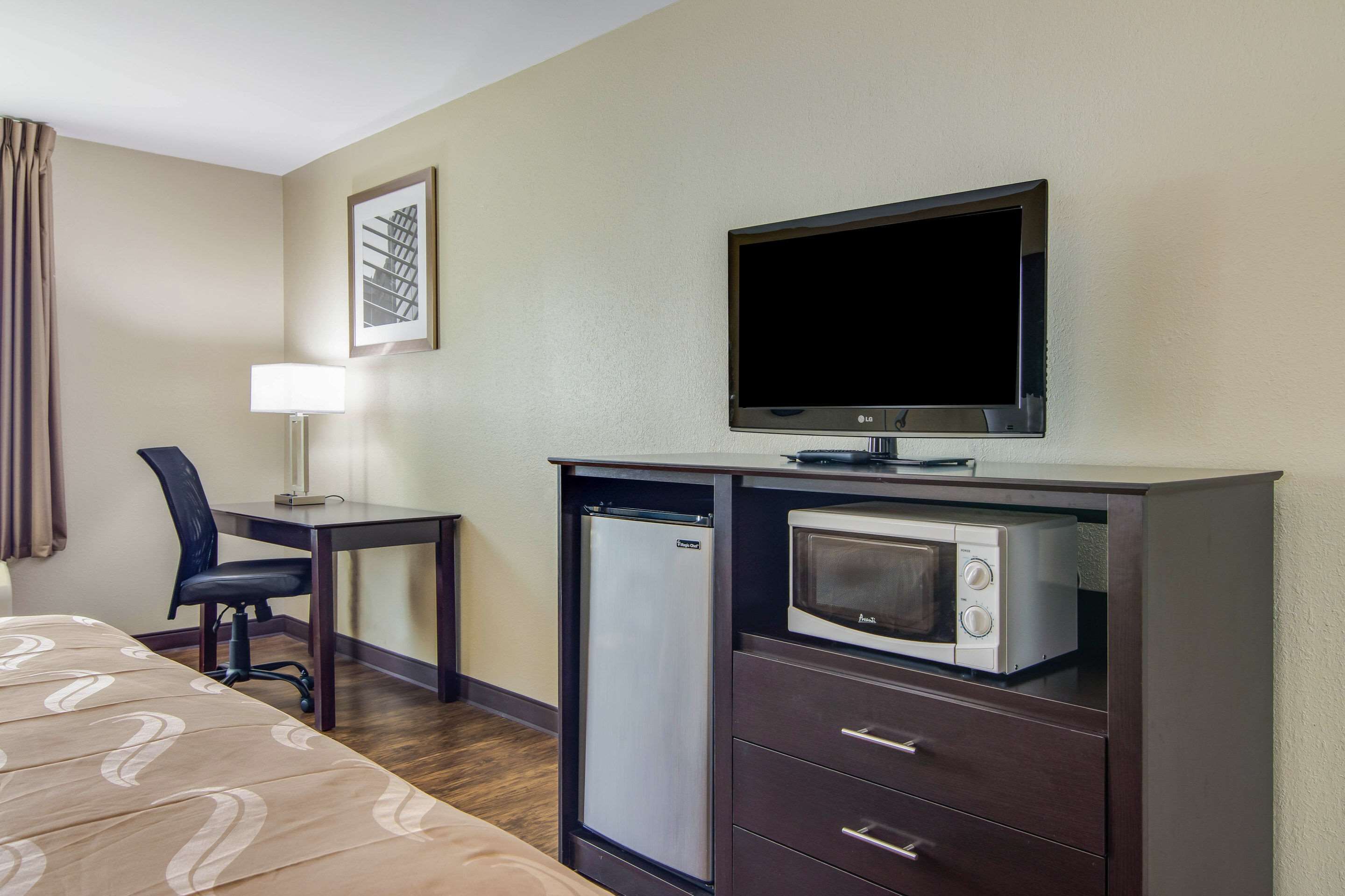 A hotel room with a bed, a desk with a chair, a TV, a microwave, and a refrigerator.