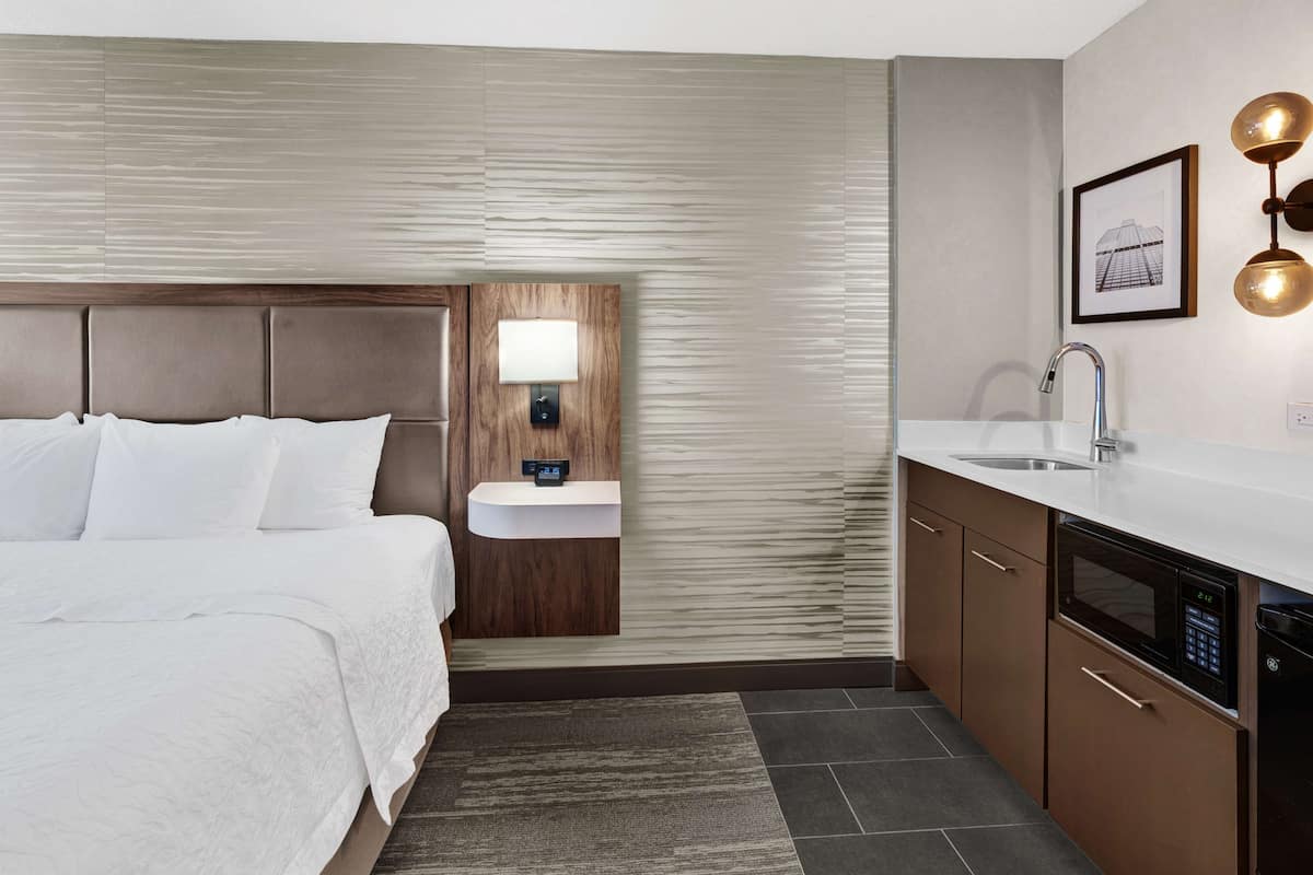 A hotel room with a bed, a bedside table with a lamp, a built-in sink, and a microwave.