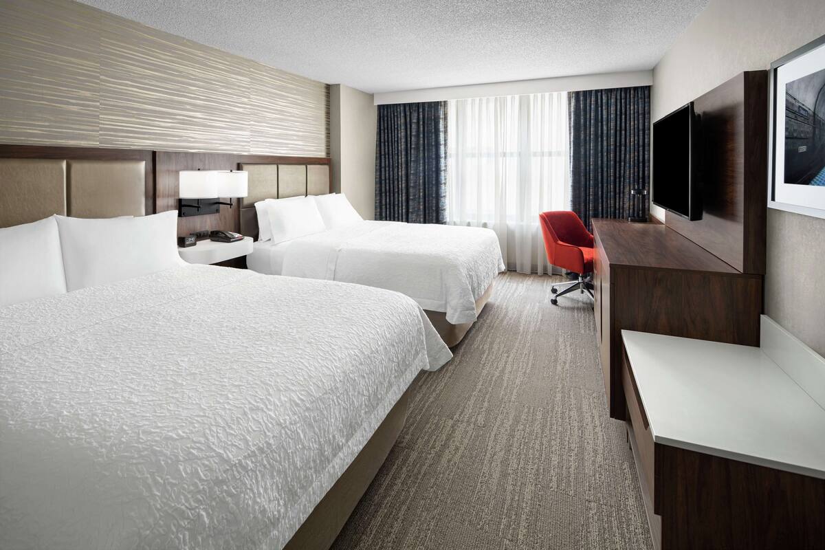 A hotel room with two beds, a desk, a chair, a TV, and a window with curtains.