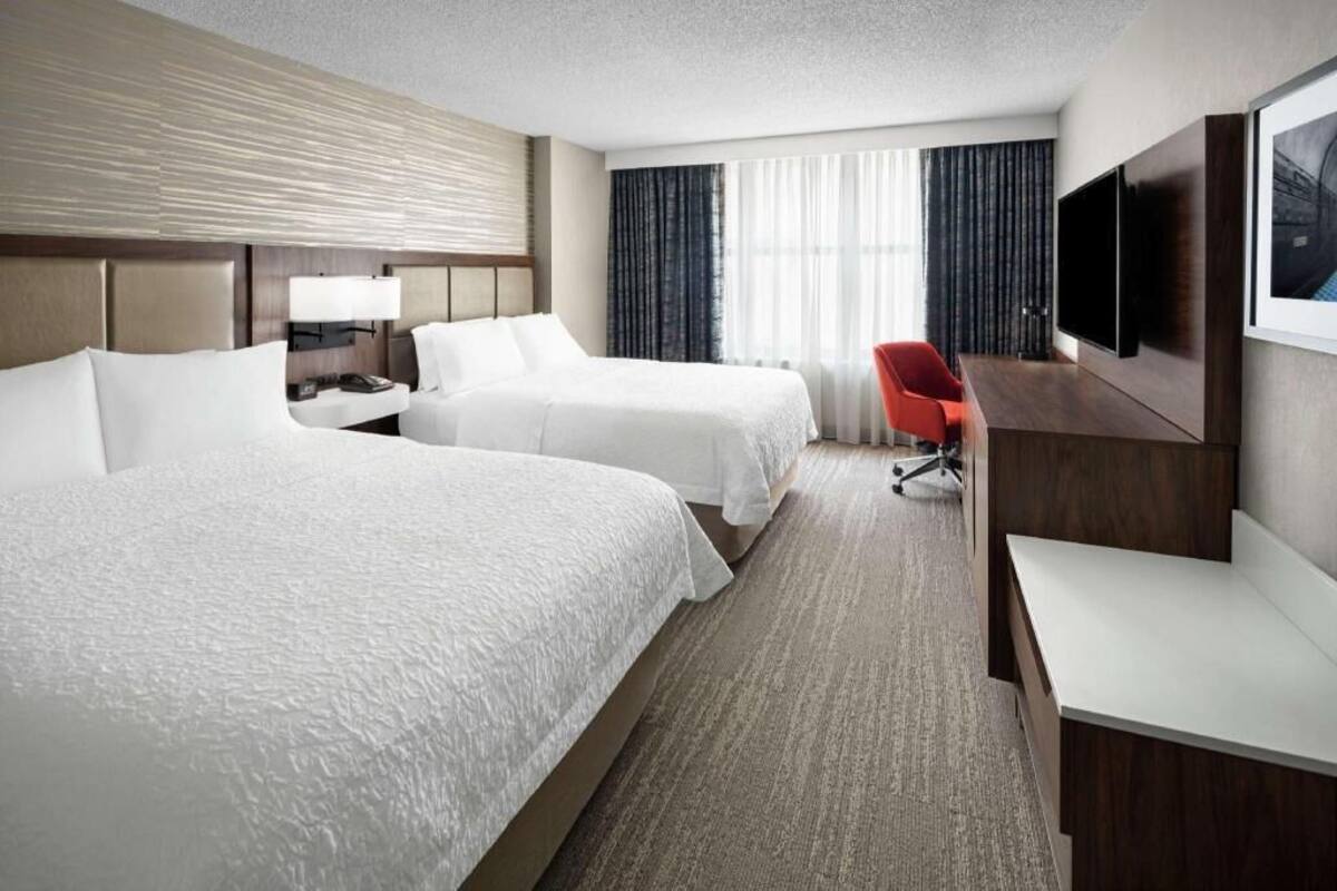 A hotel room with two beds, a desk, a chair, a TV, and a window with curtains.
