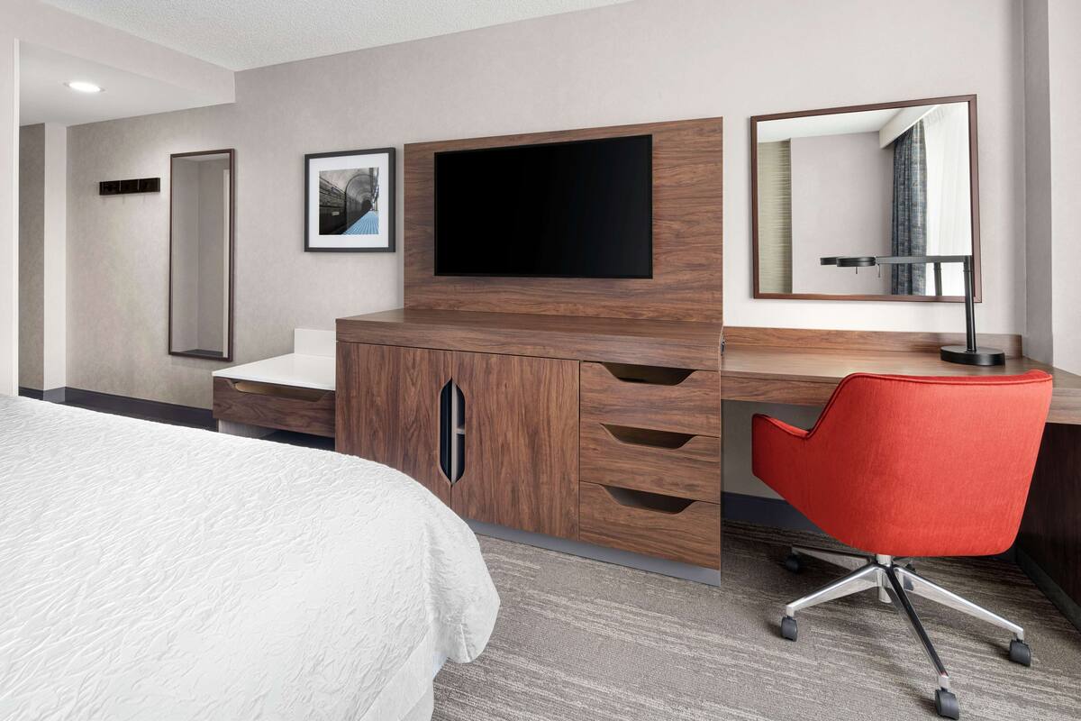 A hotel room with a bed, a wooden desk with a flat-screen TV, a red office chair, and a large mirror.
