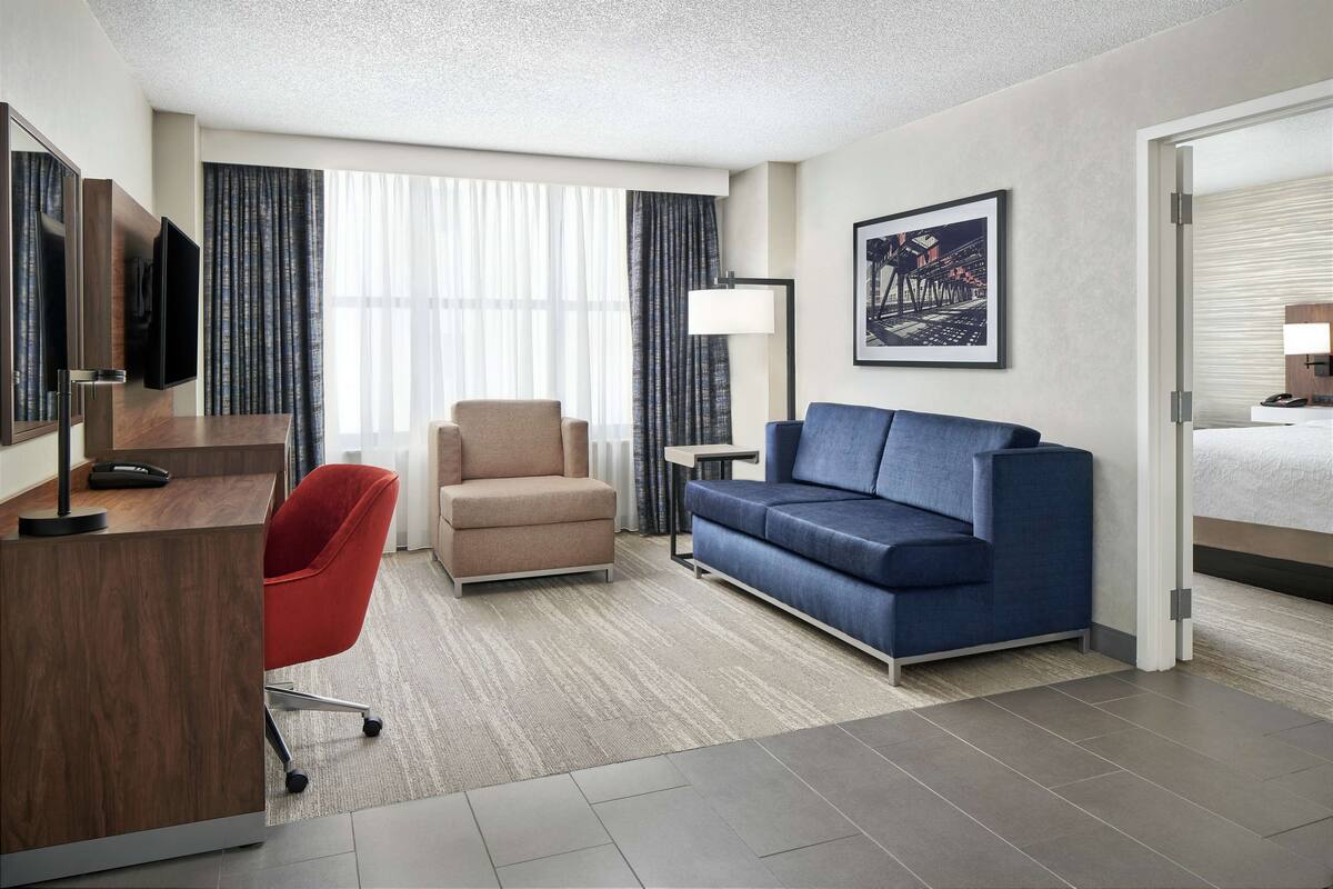 A hotel room with a desk, a red office chair, a blue sofa, a large window with curtains, a flat-screen TV, a floor lamp, and a framed picture on the wall.