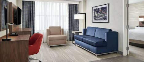 A hotel room with a desk, a red office chair, a blue sofa, a large window with curtains, a flat-screen TV, a floor lamp, and a framed picture on the wall.