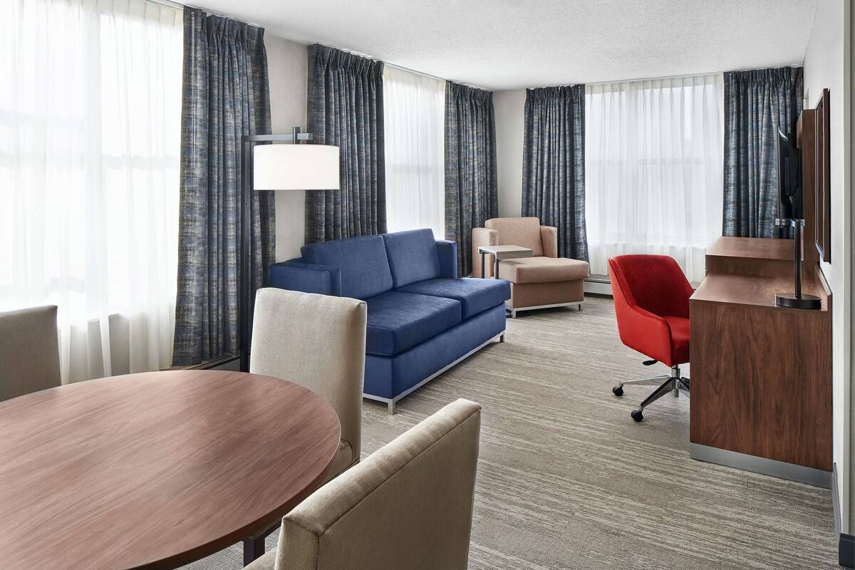 A hotel room with a blue sofa, beige armchairs, a red office chair, a wooden desk, and a TV on a wooden stand.