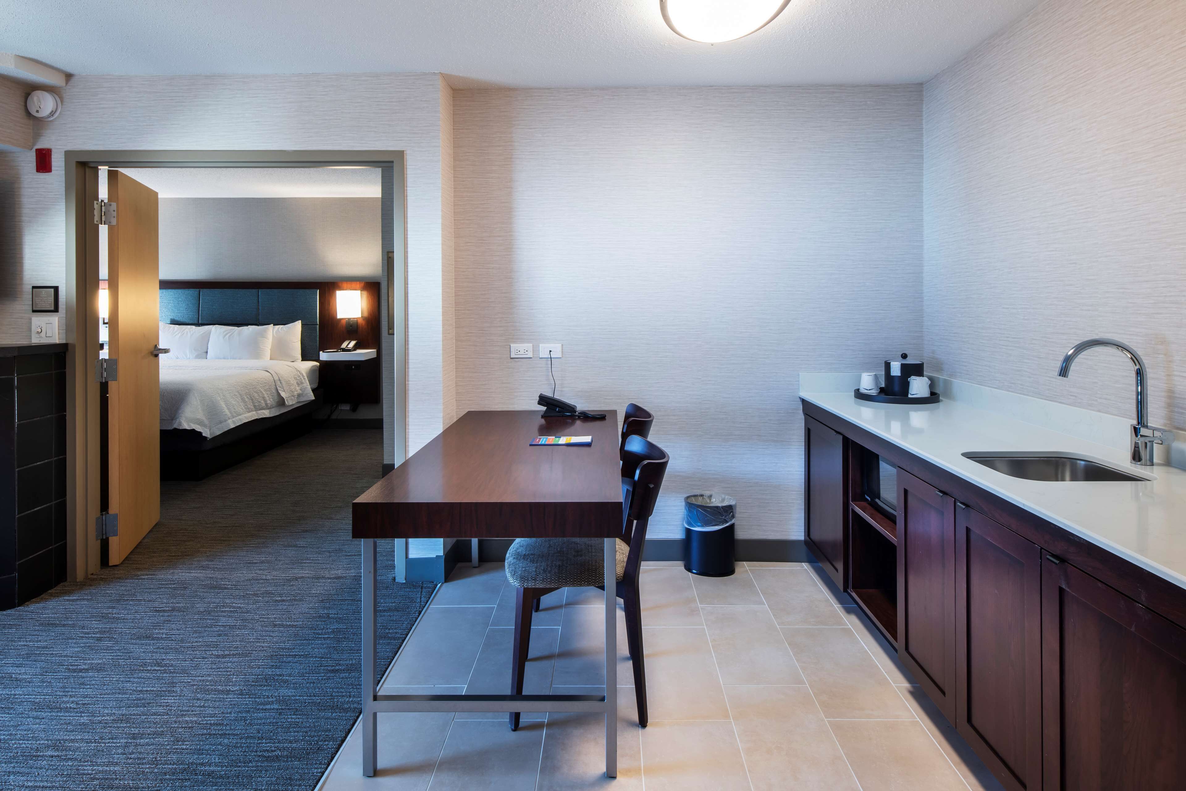 A compact hotel room with a separate dining area, a bed, a bedside table, a lamp, and a small trash bin.