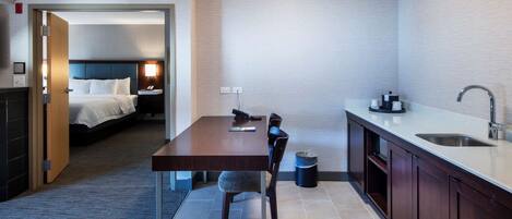 A compact hotel room with a separate dining area, a bed, a bedside table, a lamp, and a small trash bin.